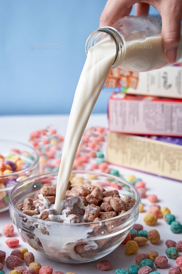 Bowl of cereal on a table, with a hand pouring milk from a glass