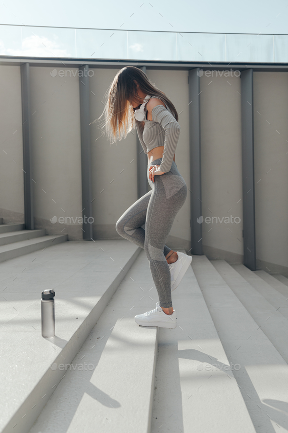 Sporty woman doing jumps and squats on stairs. Wearing grey sports active wear and headphones