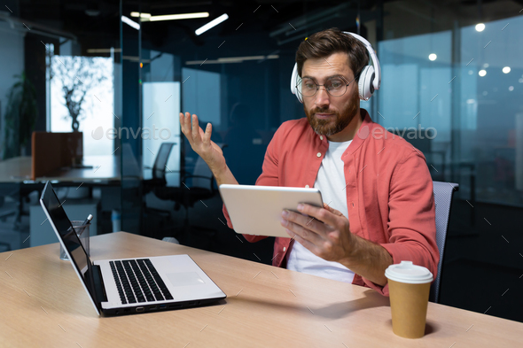 Man with headphones and tablet computer at workplace disappointed with game result, businessman