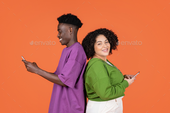 Black guy, hispanic lady standing back to back, using phones