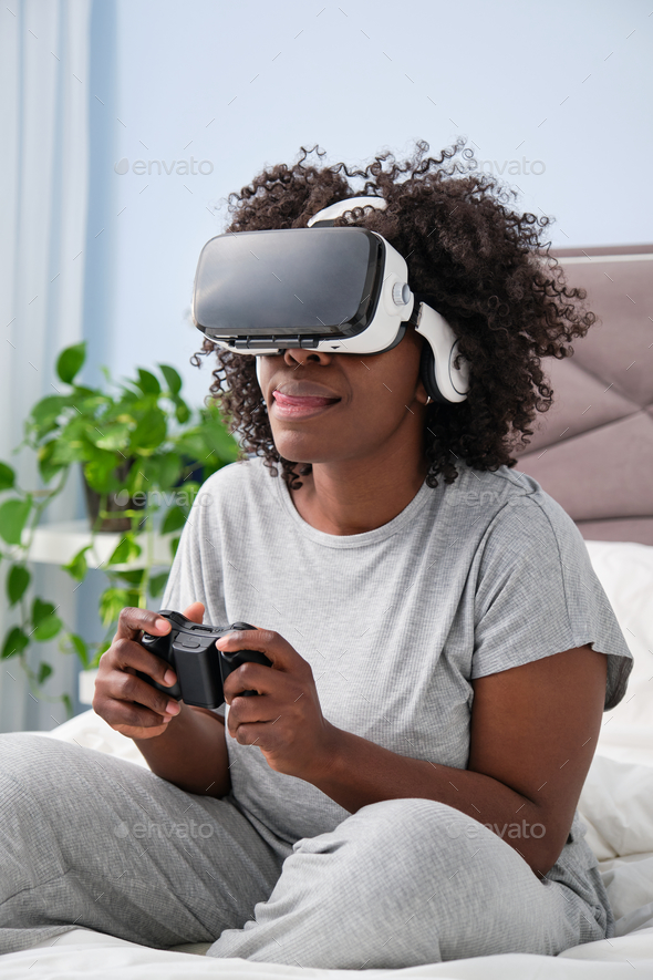 Young African woman immerses herself in virtual reality video gaming on bed.