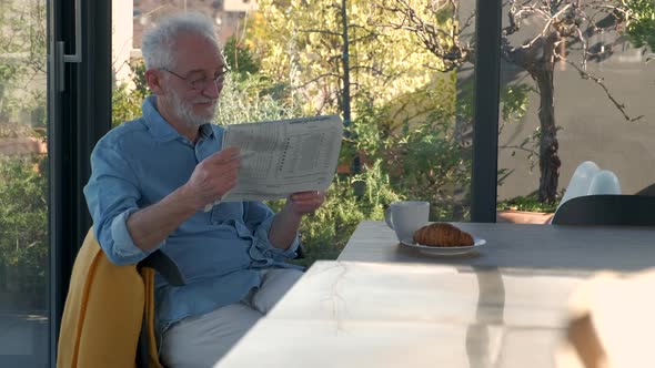 Senior man reading newspaper at breakfast table in sunny kitchen alt