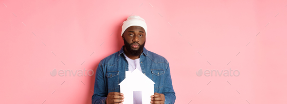 Real estate concept. Sad and tired Black man staring unamused at camera, holding paper house model