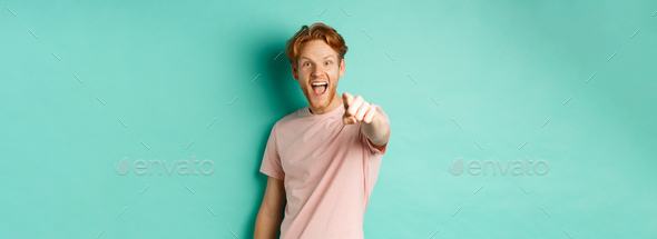 Excited young man with red hair checking out something cool, pointing finger at camera and smiling