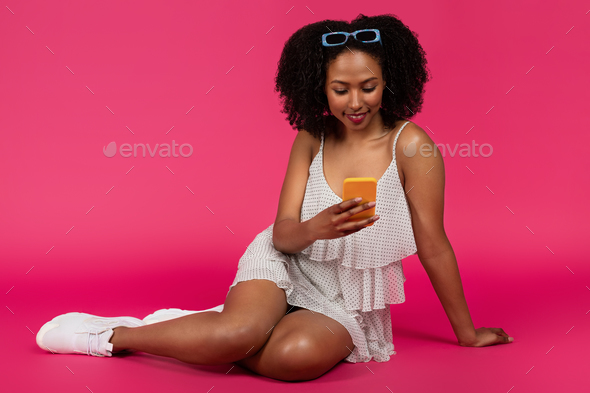Happy young black curly lady typing on smartphone, chatting in social networks, sit on floor