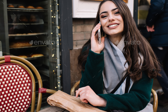 Positive woman smiling while talking on smartphone in street cafe