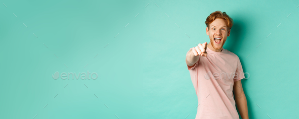 Excited young man with red hair checking out something cool, pointing finger at camera and smiling