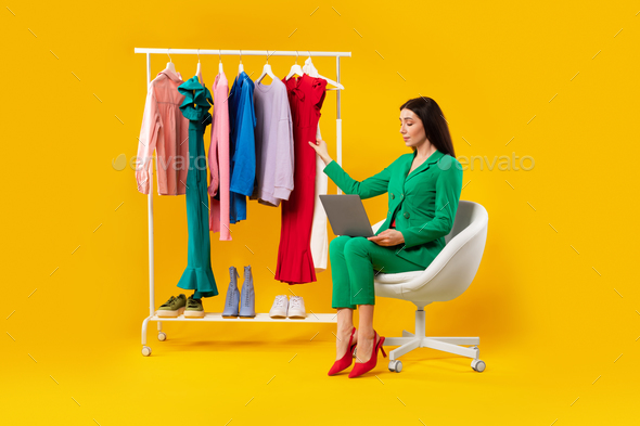 Fashion trends. Beautiful woman stylist sitting near lot of clothes on garment rack and using laptop