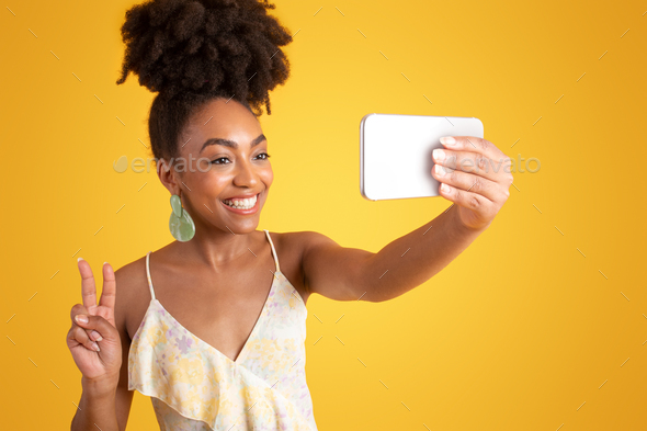 Cheerful young african american lady in dress taking selfie on smartphone, make peace sign