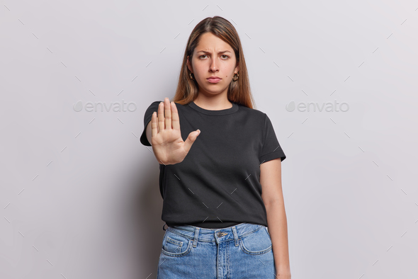Photo of serious adult European woman with dark straight hair keeps palm towards camera asks to stop