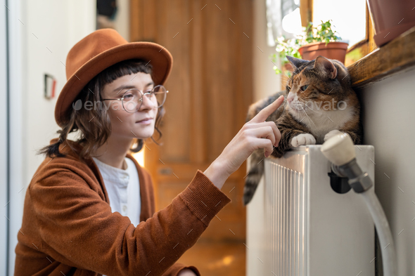 Girl in hat and glasses touches nose cat basking on batteries home, sunlight