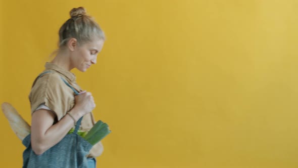 Portrait of Young Woman Walking Carrying Reusable Shopper Bag with Food on Yellow Background alt