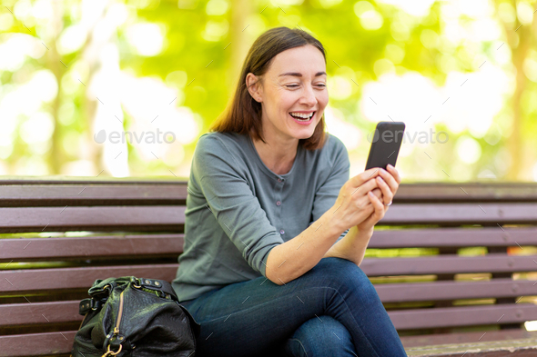 Happy Older Woman Sitting On Park Bench Looking At Cellphone Stock