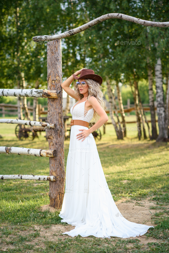 beautiful cowgirl style model posing on farmland. A beautiful girl in a white dress, a cowboy hat po