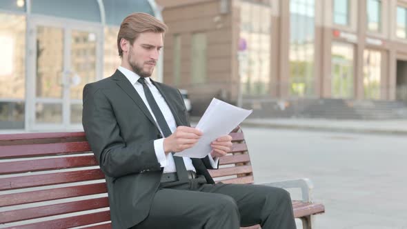 Young Businessman Reading Documents While Sitting on Bench Outdoor alt
