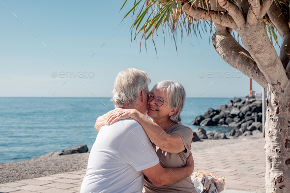 Lovely smiling senior family couple sitting on a bench close to the sea hugging with love