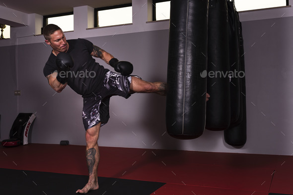 Young adult male working out by vigorously kicking a punch bag while standing on a martial arts mat