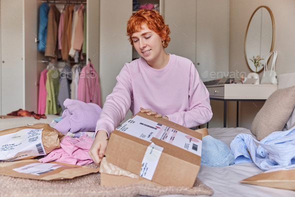 Indoor shot of redhead woman prepares delivery box sells her second hand clothes poses on bed