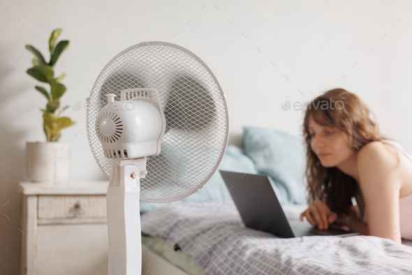 European woman cools down home with help of air conditioner fan, works with laptop in home office
