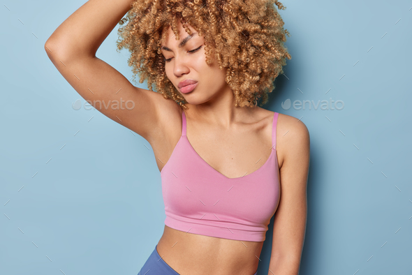 Curly haired woman dressed in cropped top smells her sweaty armpit after workout frowns face