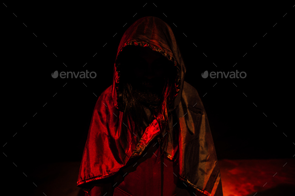 Mysterious figure in a hooded cloak illuminated by red lights in a dark room