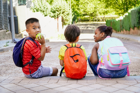 multiethnic kids with backpacks sitting on the street at school entrance eating apples.