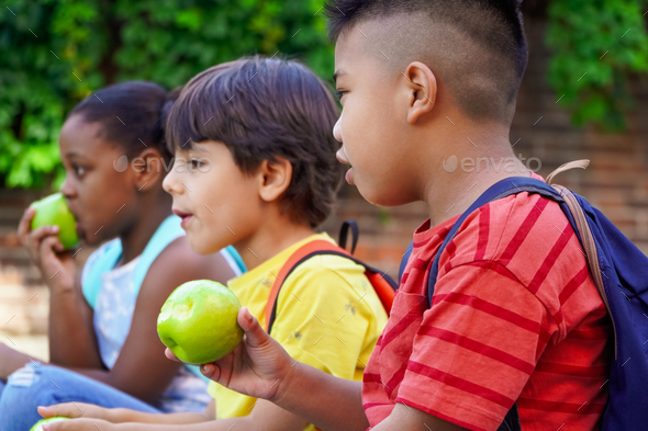 multiethnic kids with backpacks sitting on the street at school entrance eating apples.