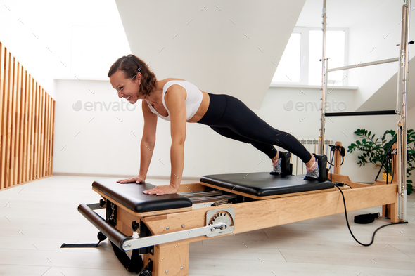 A woman is doing Pilates on a reformer bed in a bright studio. A slender,  flexible brunette in a Stock Photo by Gerain0812
