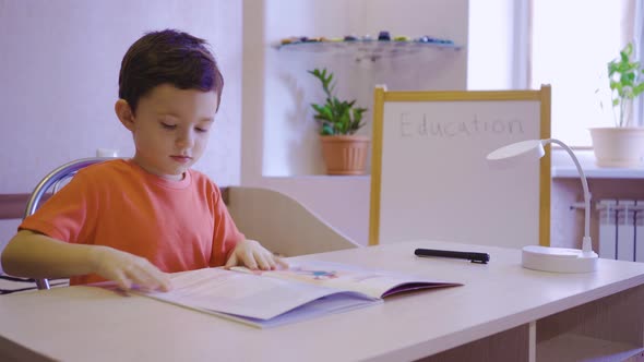 A Little Brunette Boy Reading a Book While Sitting at Home at a Table