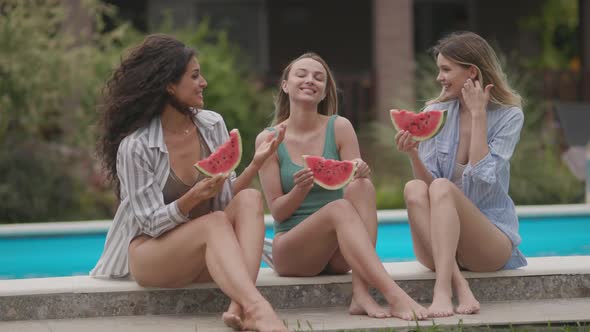 Three cute young women sitting on by the swimming pool and eating watermelon in the house backyard