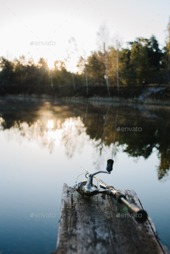 Fishing rod on a bridge on pond background. Spinning with reel on pier river.  Misty morning on lake Stock Photo by kurinchukolha