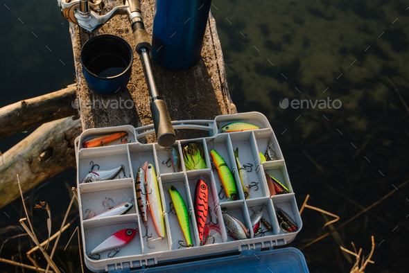Fishing tackle - fishing spinning, rod, reel, hooks, fly, bait, lures in  box on wooden pier on pond Stock Photo by kurinchukolha