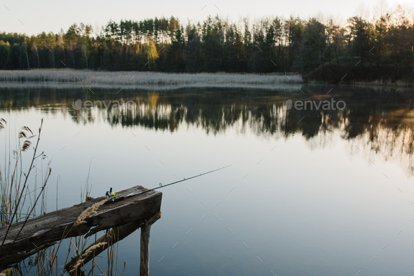Fishing rod, spinning reel on the background pier river bank. Sunrise. Fog  against the backdrop Stock Photo by kurinchukolha