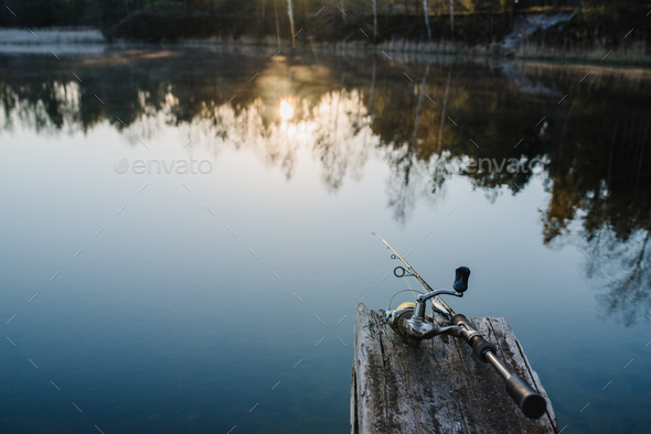 Fishing rod, spinning reel on the background pier river bank. Sunrise. Fog  against the backdrop Stock Photo by kurinchukolha