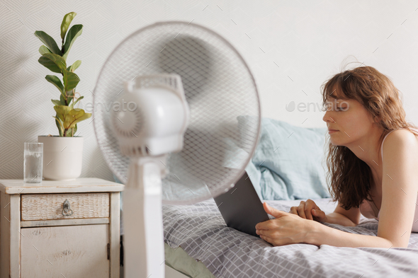 European woman cools down home with help of air conditioner fan, works with laptop in home office