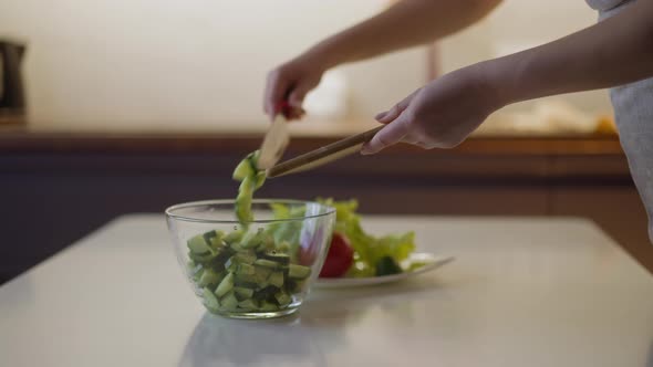 Woman Puts Cucumber Slices Into Glass Bowl on Table alt
