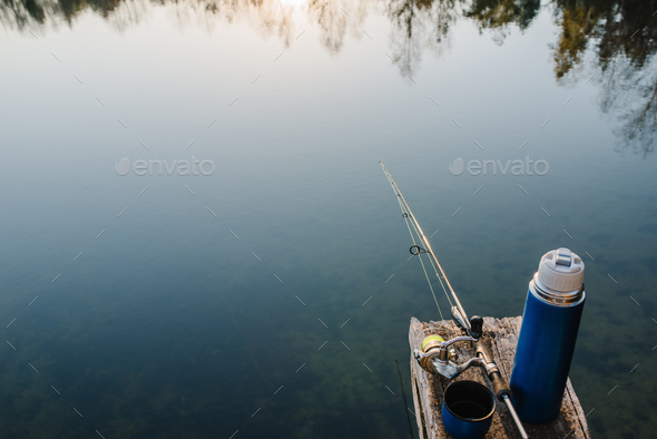 Fishing rod on a bridge on pond background. Spinning with reel on pier  river. Thermos with coffee, Stock Photo by kurinchukolha