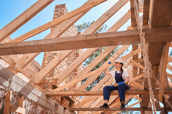 Responsible woman general contractor sitting on beam of roof frame