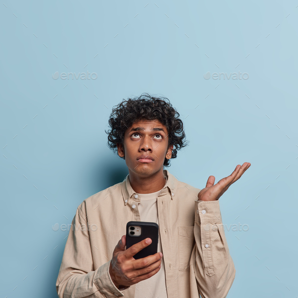 Vertical shot of hesitant curly haired man keeps palm raised up and feels clueless takes decision