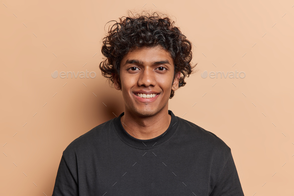 Portrait of gentle cute woman with curly hair keeps hands on cheeks smiles  pleasantly looks directly at camera wears casual shirt poses against pink  background. Human face expressions concept Stock Photo |