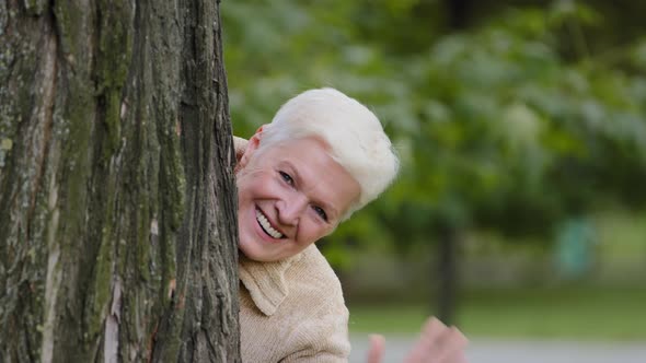 Funny Cheerful Mature Middle Aged Woman Hiding Behind Tree Plays Having Fun Peeping Out Waving Hand alt