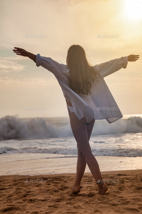 From behind young woman in white shirt going with hands to side at ocean