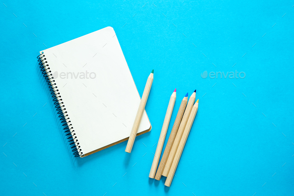 Blank sketchbook with colored pencils on blue background, top view