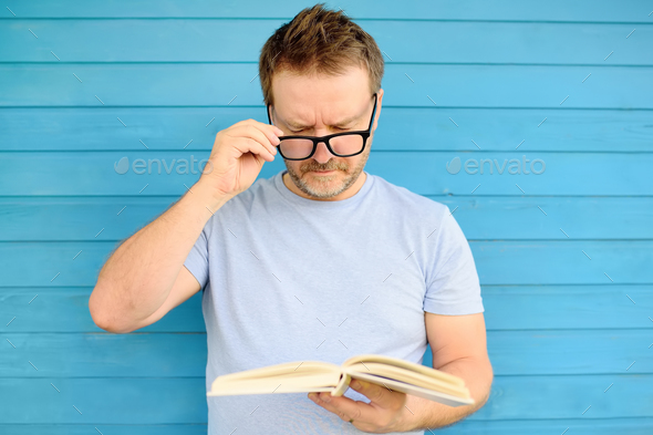 Portrait of mature man with big black eye glasses trying to read book but having difficulties seeing