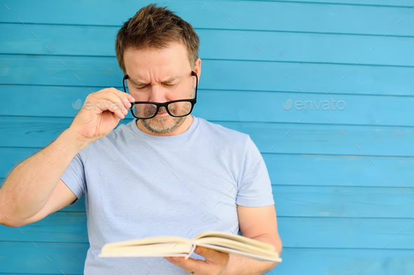 Portrait of mature man with big black eye glasses trying to read book but having difficulties seeing