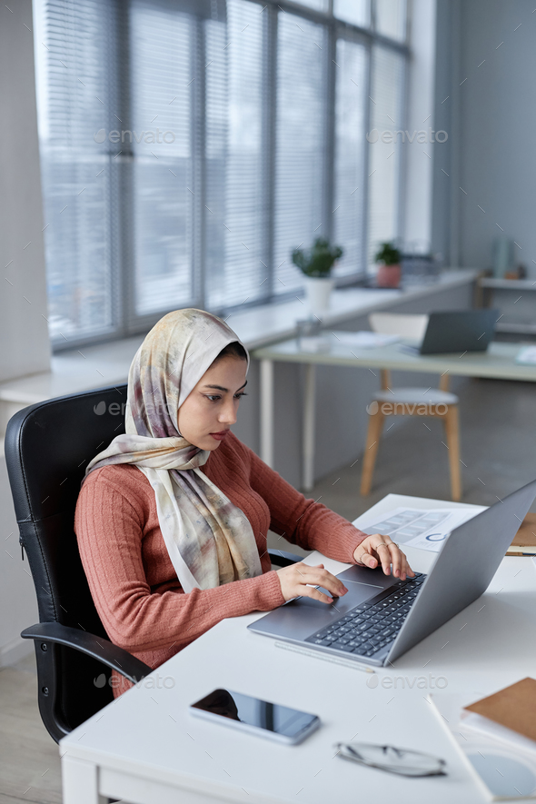 Young serious female analyst in headscarf using laptop while working