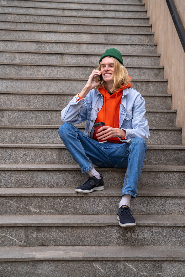 Happy hipster guy millennial talking over phone while sitting on pedestrian crossing stairs