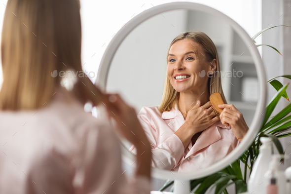 Smiling Middle Aged Blonde Lady Brushing Her Hair With Comb Near Mirror
