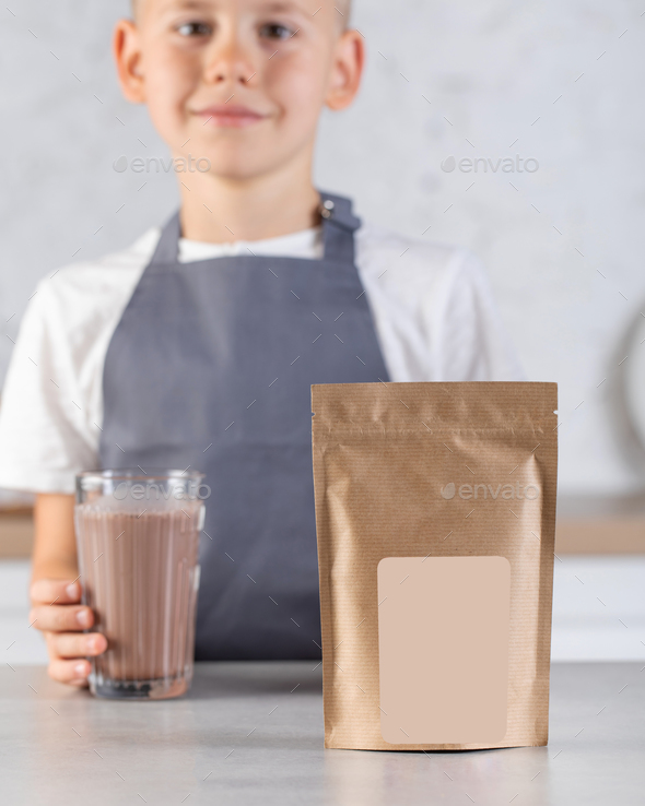 A cute joyful boy holds a glass of cocoa and a craft package with a place for a logo. Cocoa powder