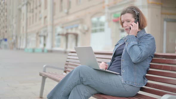 Angry Old Woman Talking on Phone and Using Laptop While Sitting Outdoor on Bench alt